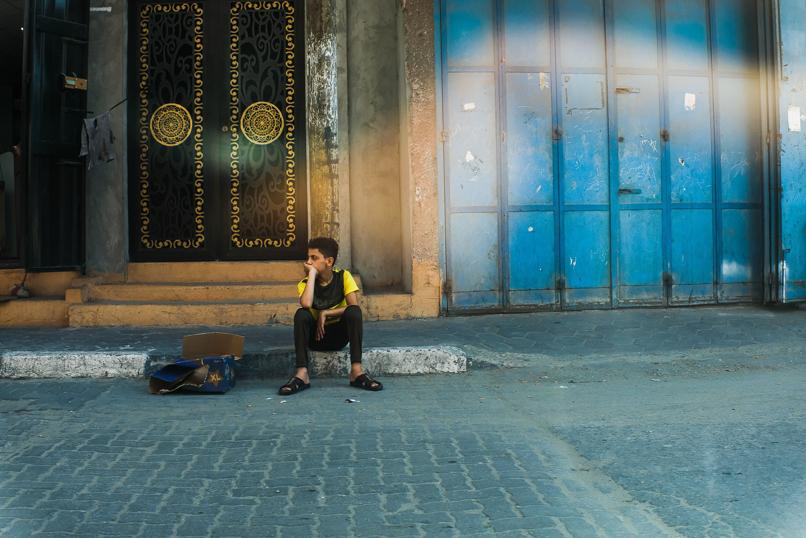 Vaikinas, sėdintis ant šaligatvio ilgiausioje „Saladino” gatvėje. Jabalia, Gaza. 2021 m. rugpjūčio 2 d. Nuotrauka: Mahmoud Nasser.